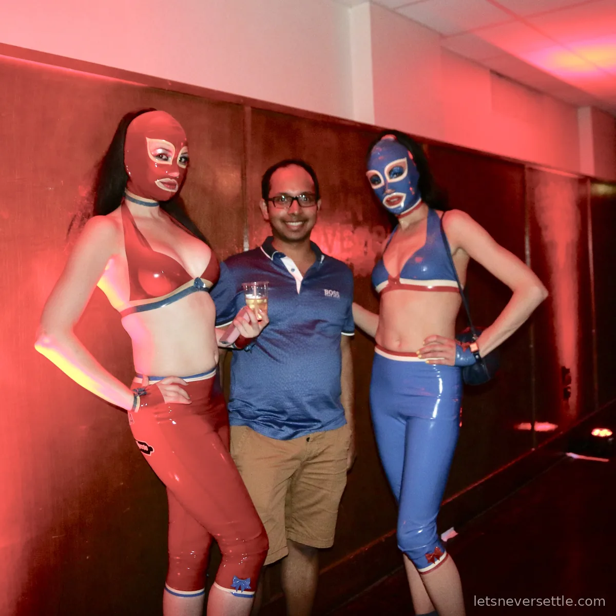 Dileep holding a drink and standing between two masked luchadoras in red and blue costumes in a red lit corridor