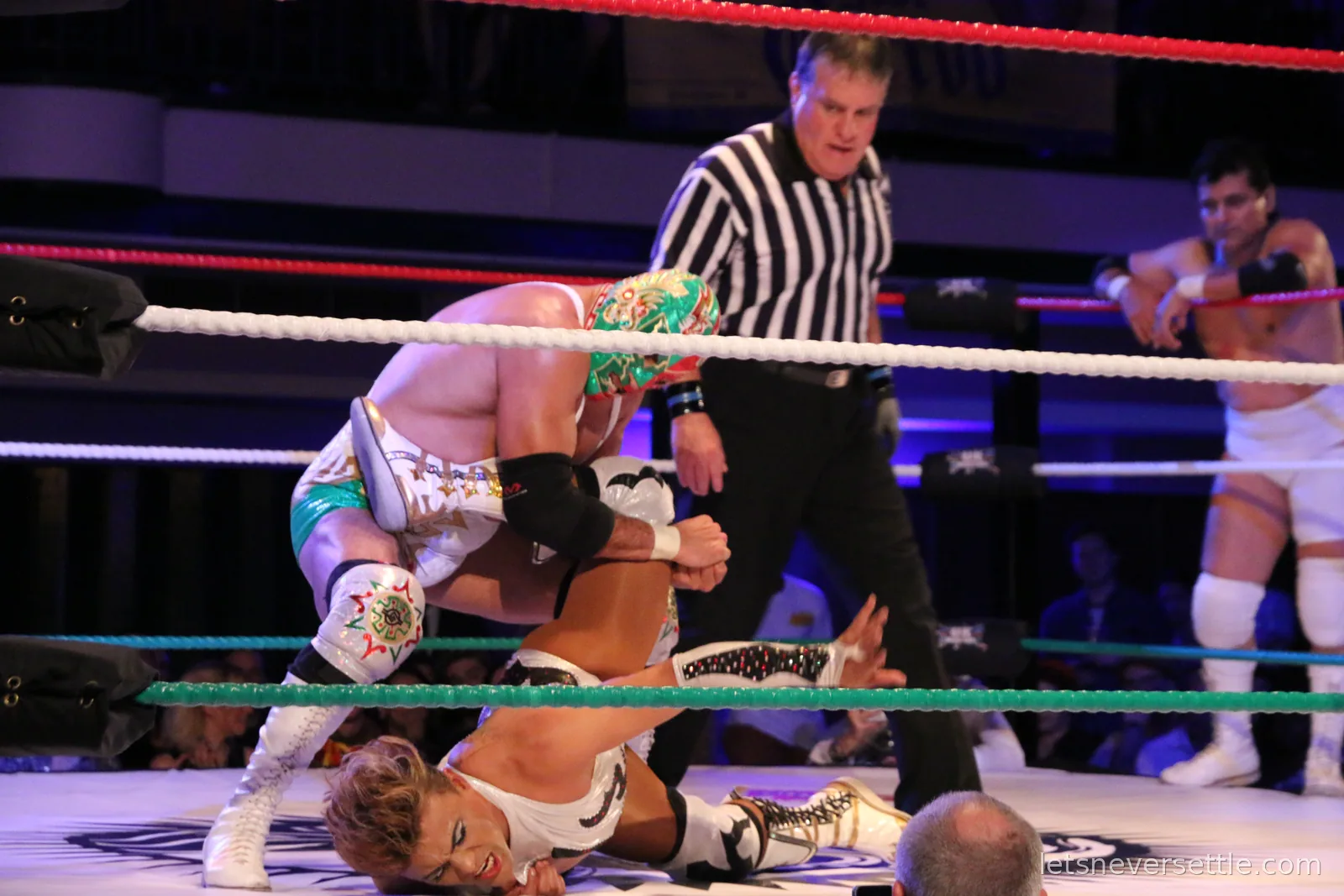 A referee in a striped shirt crouching low to count a pin while one masked wrestler holds another down on the canvas