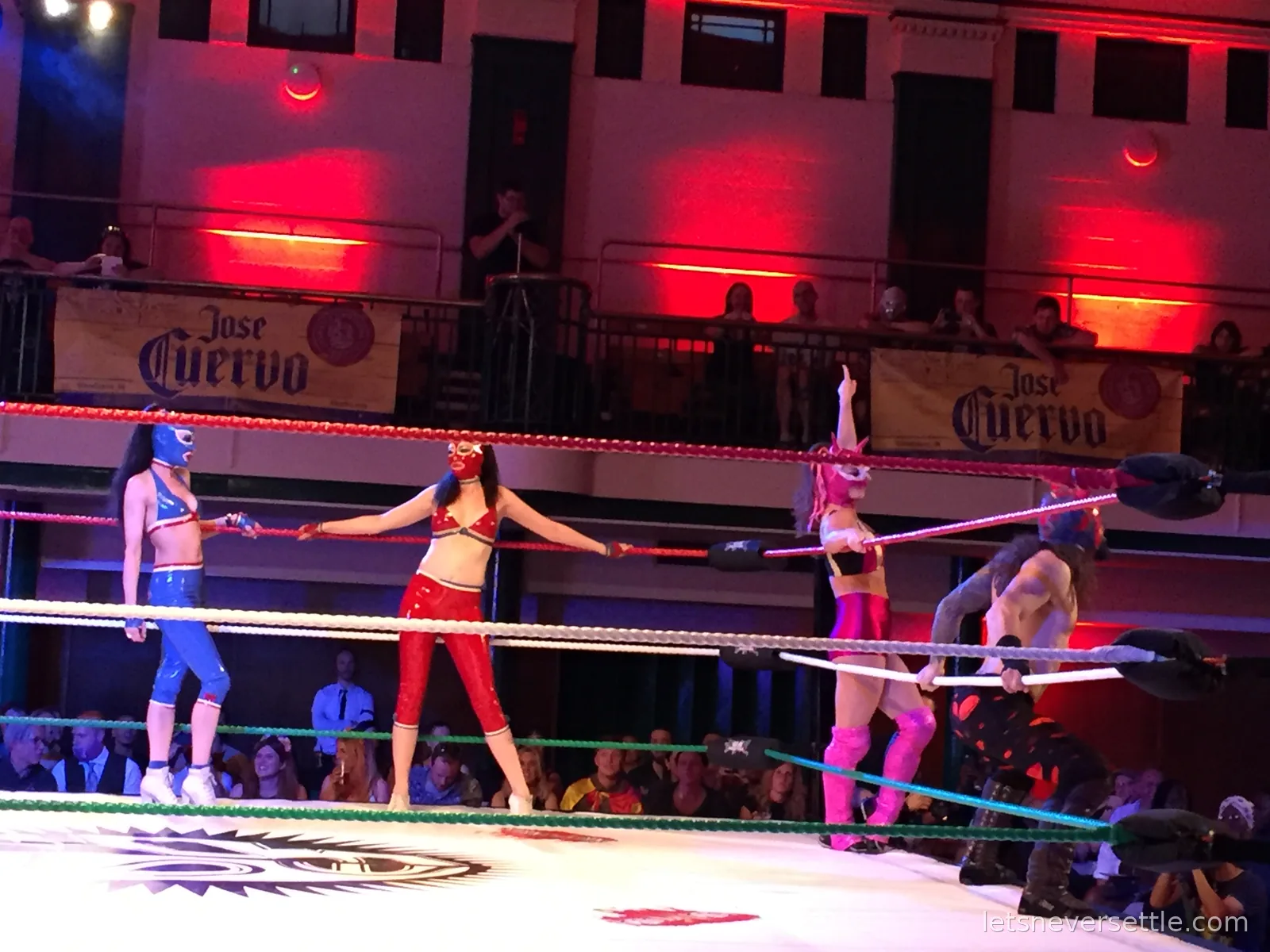 Three masked luchadoras in blue, red and pink costumes squaring up in the ring under red lights, a Jose Cuervo banner behind