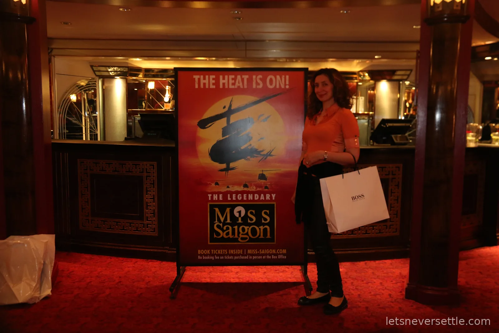 Inga standing beside a Miss Saigon poster reading The Heat Is On in the red art deco foyer of the theatre, holding a paper shopping bag