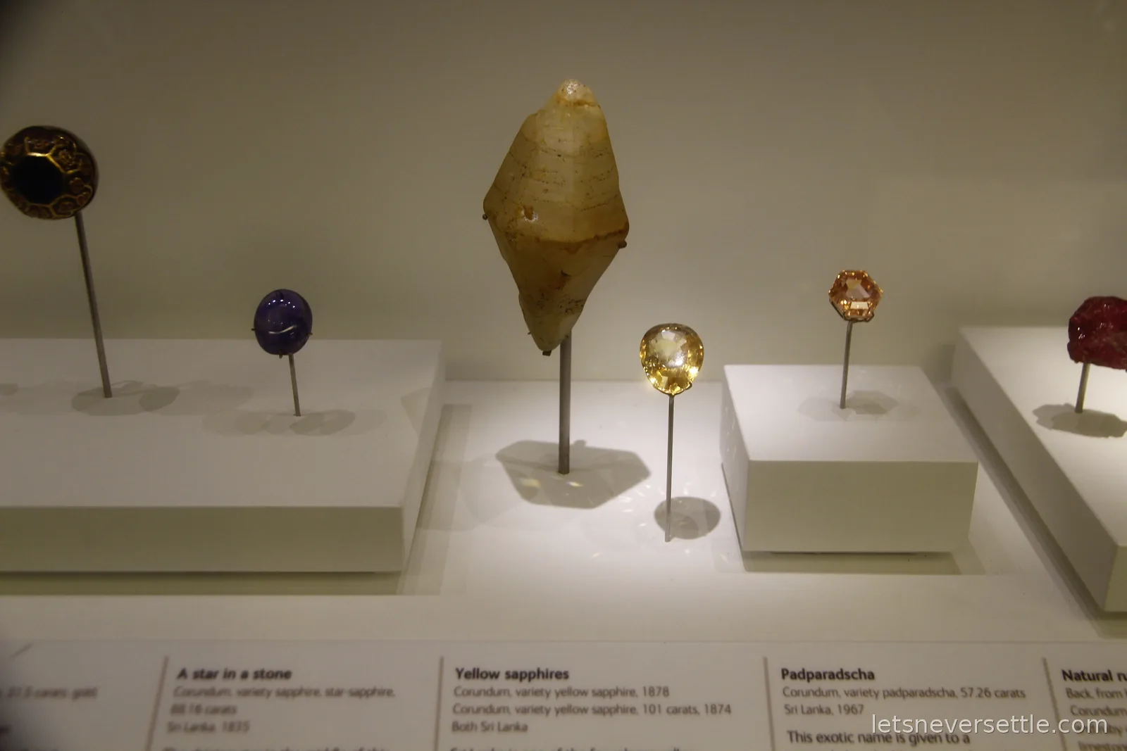 Cut sapphires mounted on thin stands in a museum case, with labels reading yellow sapphires and padparadscha from Sri Lanka