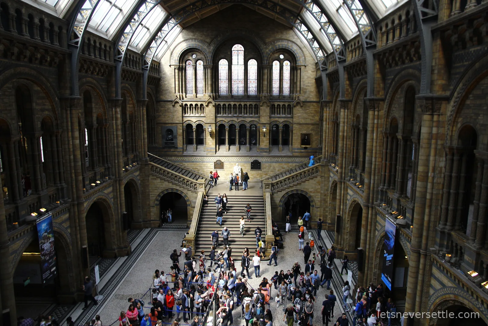 The central hall of the Natural History Museum seen from an upper balcony, crowds on the wide stone staircase beneath vaulted terracotta arches and tall windows