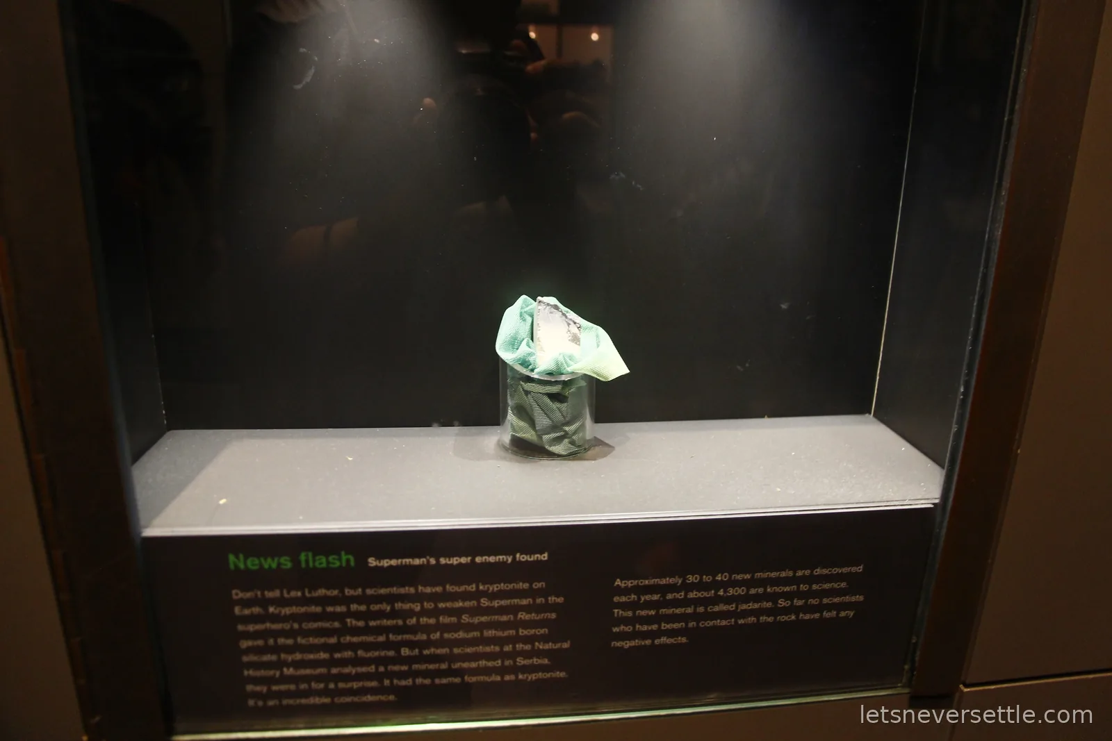 A small grey mineral sample in a dark museum case below a label headed News flash, Superman's super enemy found