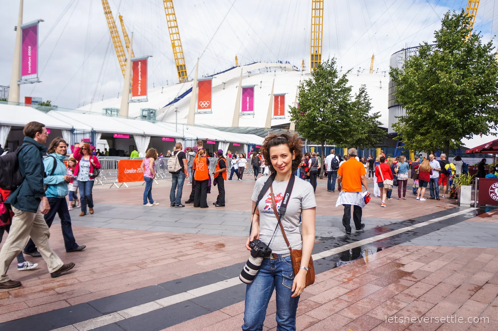 Inga in a London 2012 t-shirt with a Canon camera outside the O2, its white tented roof and yellow masts behind her, hung with pink London 2012 and Olympic rings banners