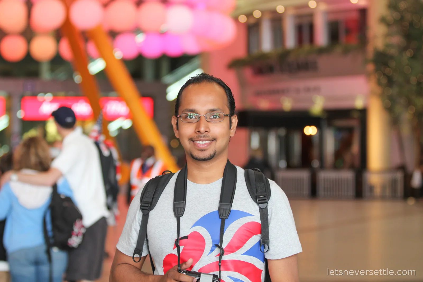 Close portrait of Dileep in a Union Jack lion t-shirt on the O2 concourse, the hanging pink and orange ball sculpture blurred behind him
