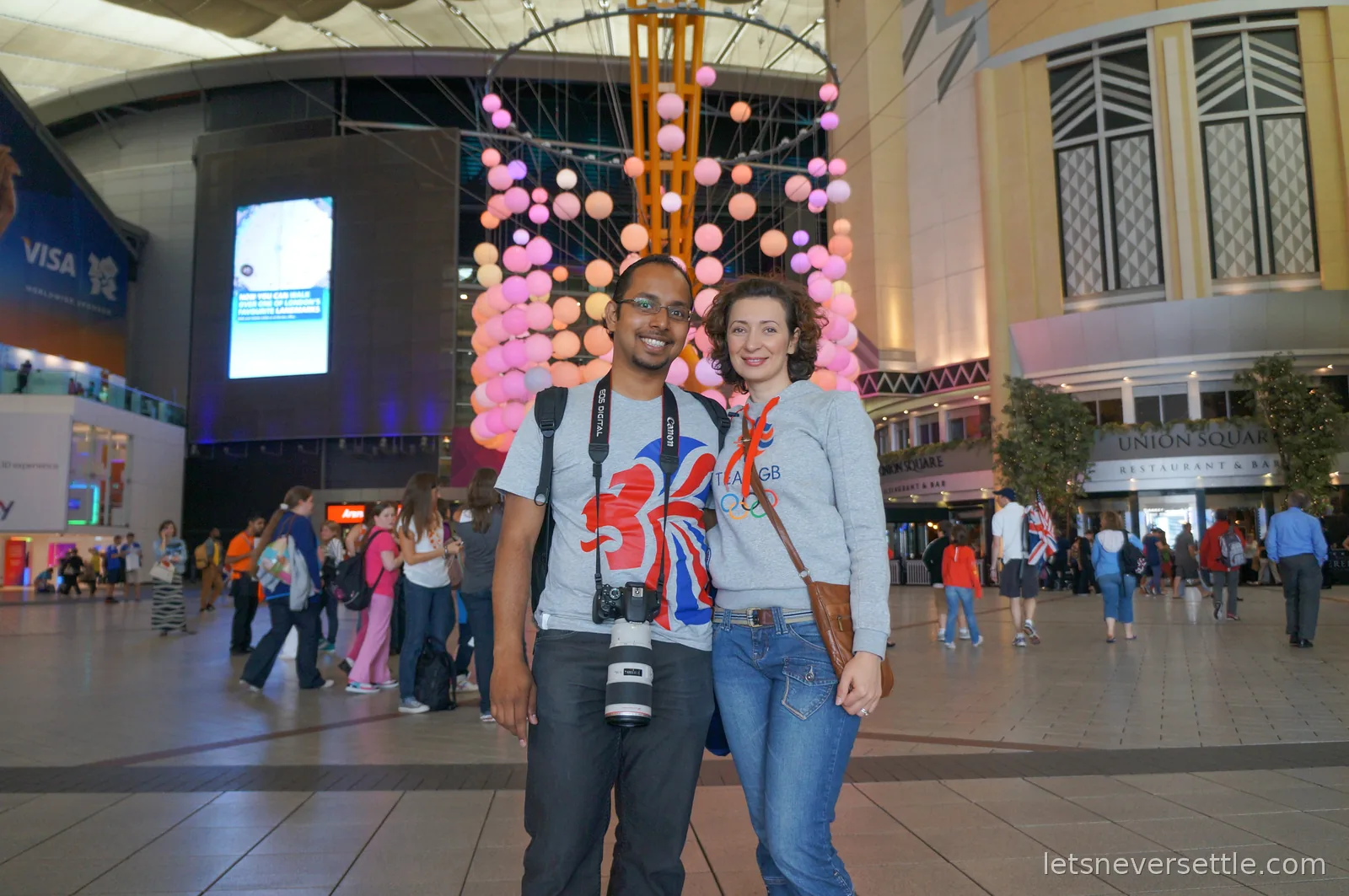 Dileep in a Union Jack lion t-shirt and Inga in a Team GB sweatshirt standing under a hanging sculpture of pink and orange balls inside the O2, a Union Square Restaurant and Bar sign to the right