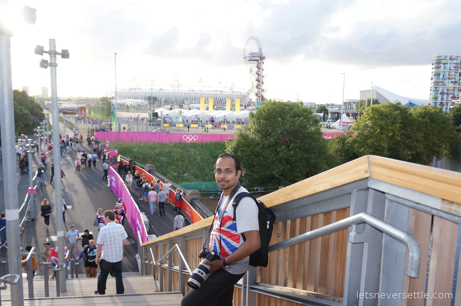 Dileep in a Union Jack t-shirt on a staircase in the Olympic Park, with the Olympic Stadium, the red Orbit tower and the rings on a pink banner behind