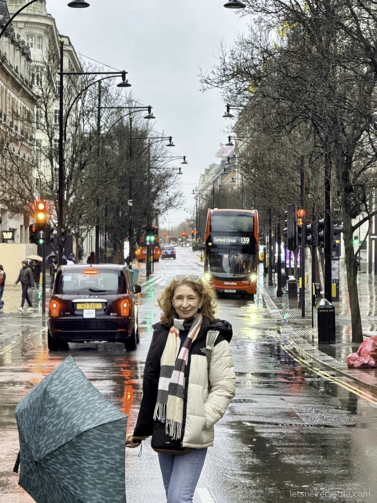 Inga holding an umbrella on a rainy Oxford Street with a black cab and a red number 139 double-decker bus behind her