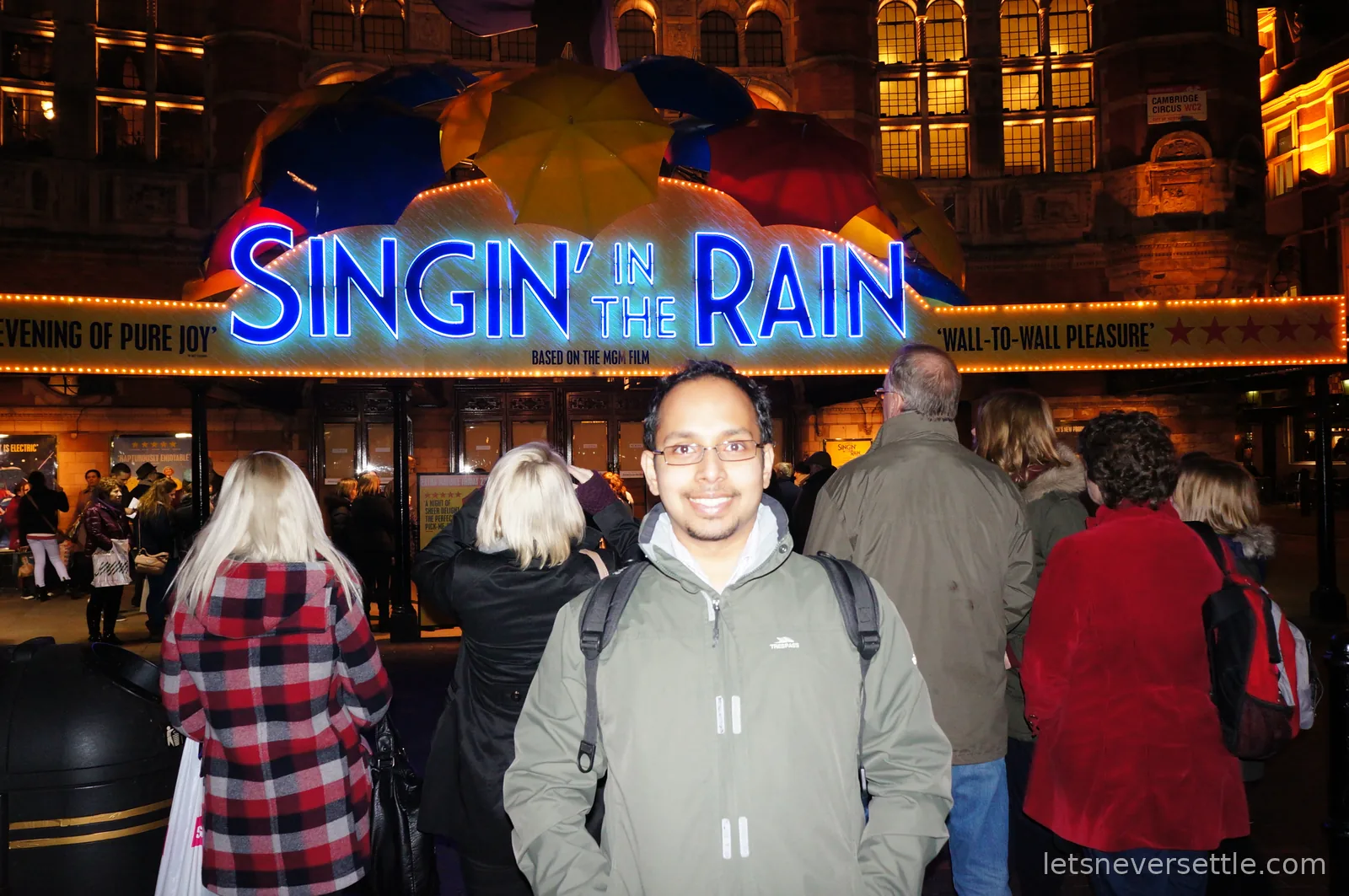 Dileep in a green coat outside the Palace Theatre at night, under the Singin' in the Rain marquee topped with colourful umbrellas