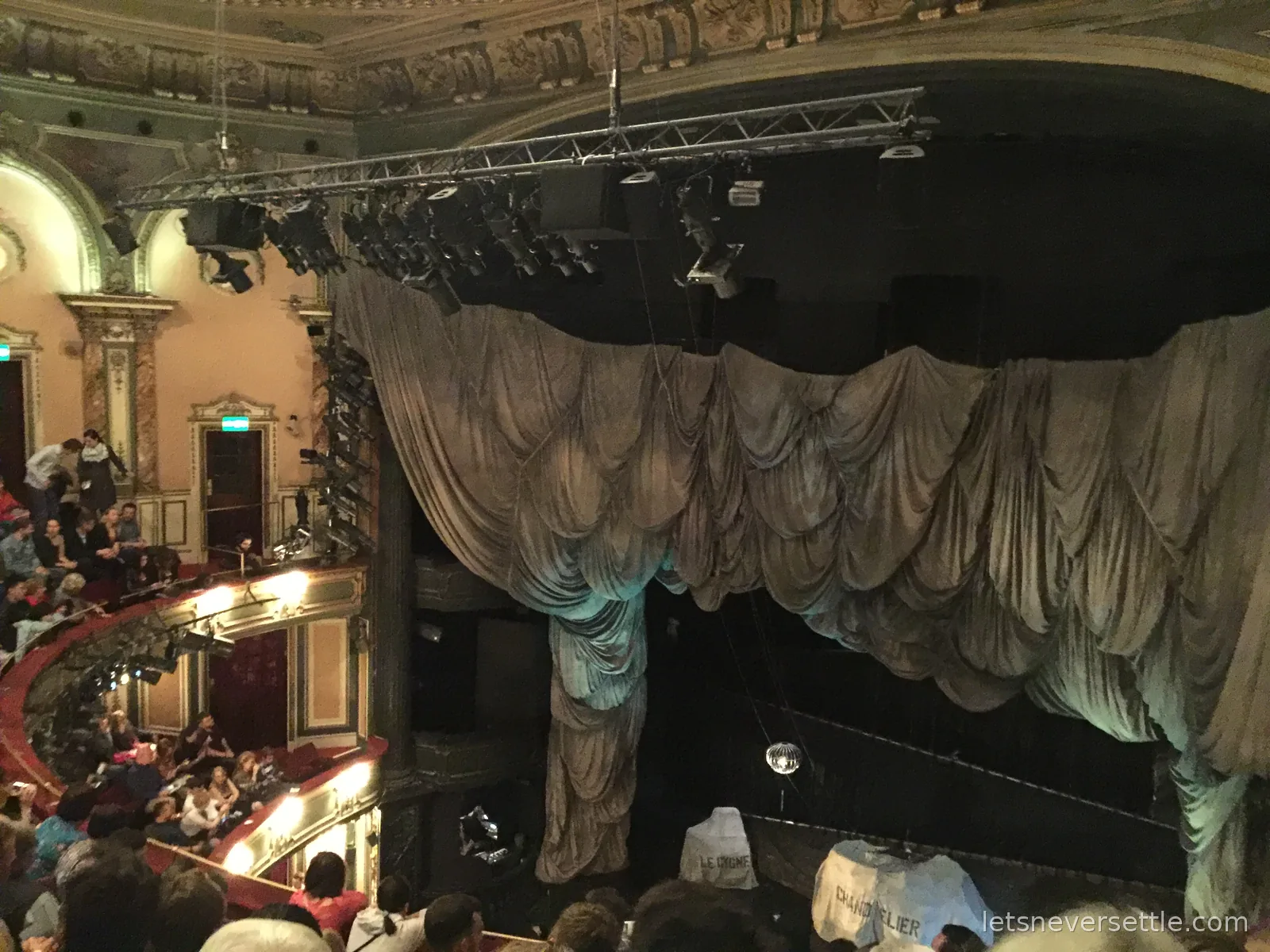 A West End auditorium filling up before The Phantom of the Opera, the stage hidden behind a huge draped grey curtain under the lighting rig