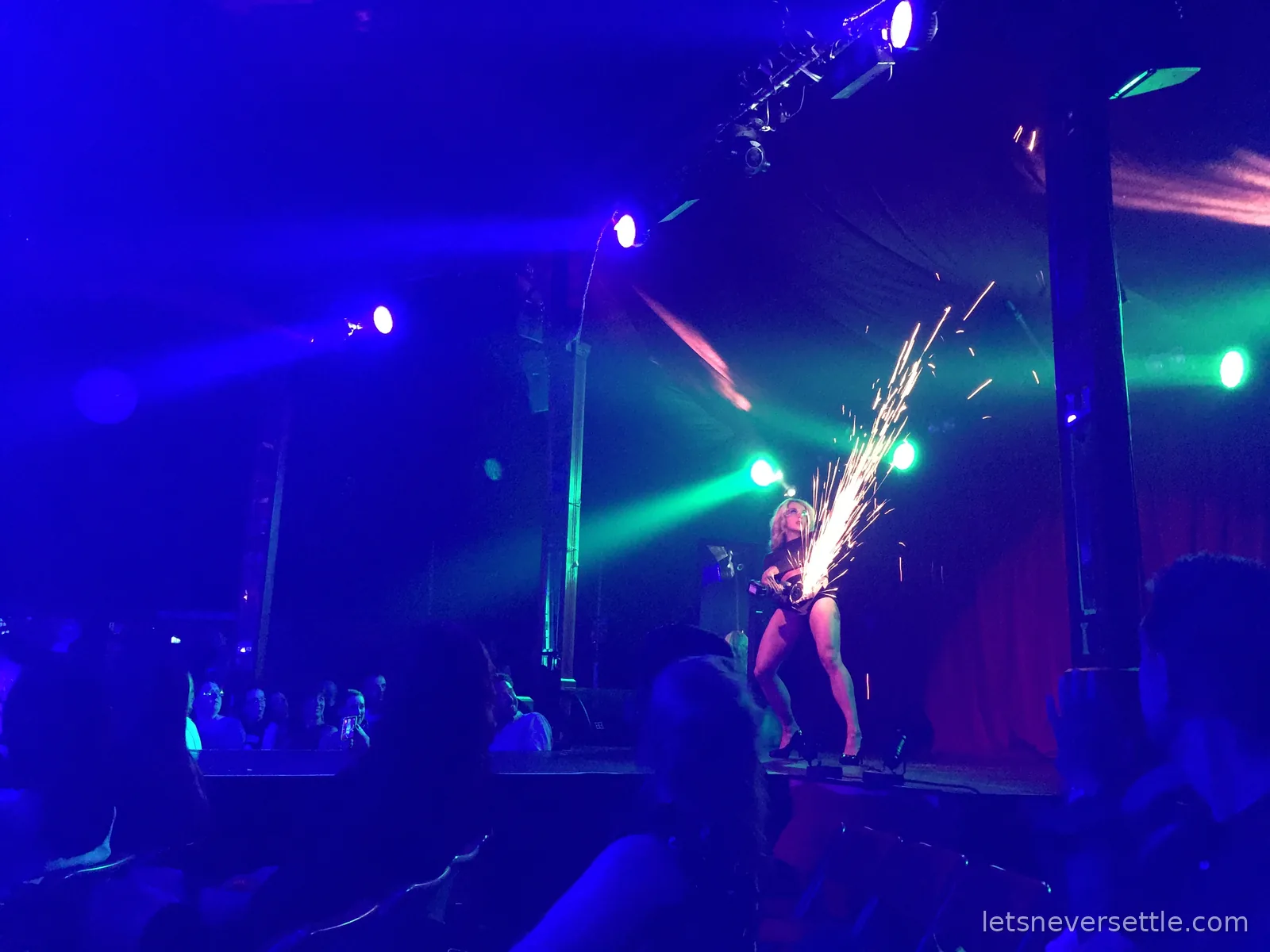 A performer on a spiegeltent stage sending a fountain of sparks from an angle grinder under blue and green stage lights