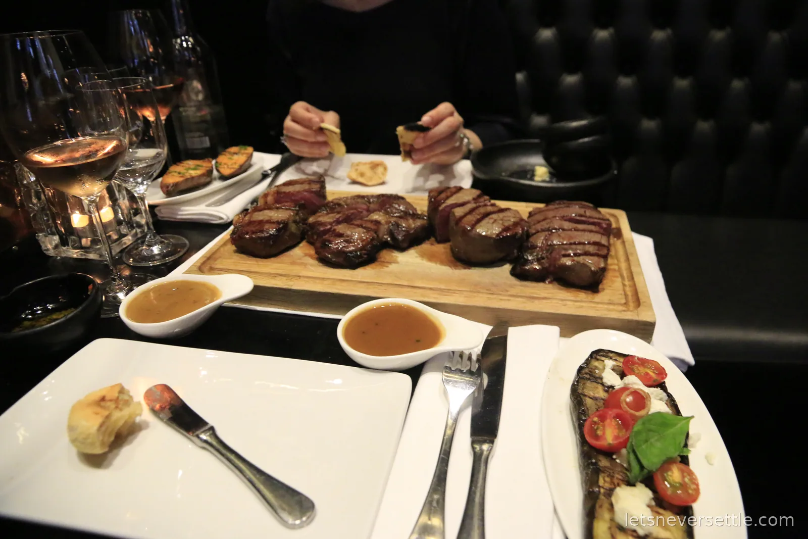 A wooden board of sliced steaks with gravy boats, grilled vegetables and wine glasses on a dark restaurant table