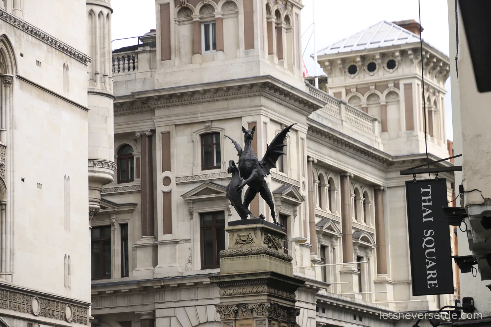 A black winged dragon statue on a stone plinth marking the City of London boundary, the Royal Courts of Justice rising behind it