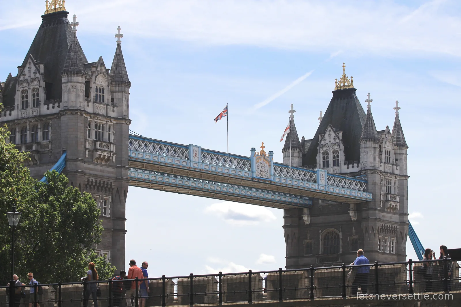 The twin Gothic towers and pale blue walkways of Tower Bridge under a summer sky, a Union Jack flying between them