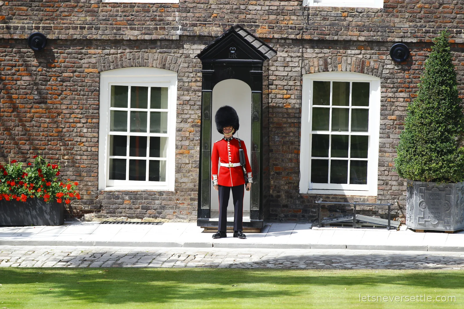 A Queen's Guard soldier in red tunic and bearskin standing at his sentry box against the brick wall at the Tower of London, red geraniums to one side