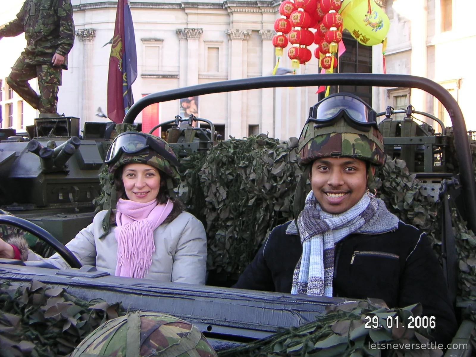 Inga and Dileep wearing camouflage helmets with goggles while sitting in an open army vehicle at Trafalgar Square, red lanterns strung overhead
