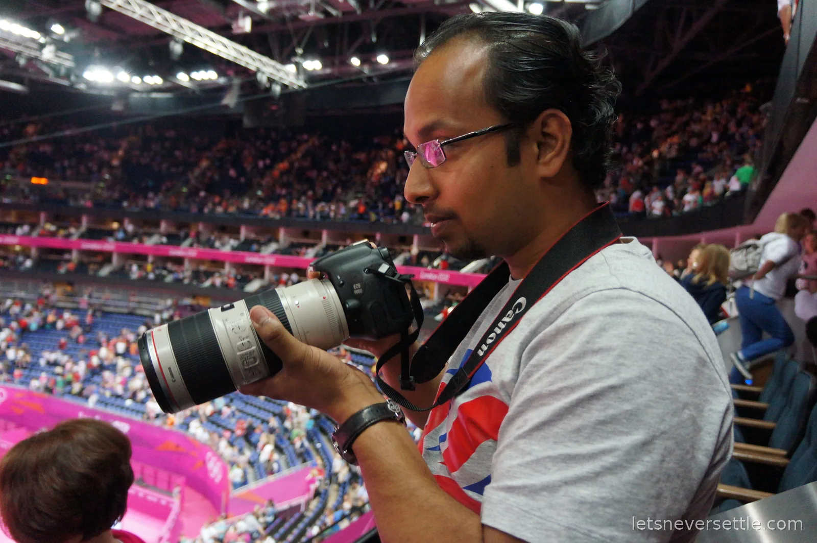 Dileep in a Union Jack t-shirt holding a Canon EOS 600D with a long white telephoto lens, looking down at the arena bowl full of spectators