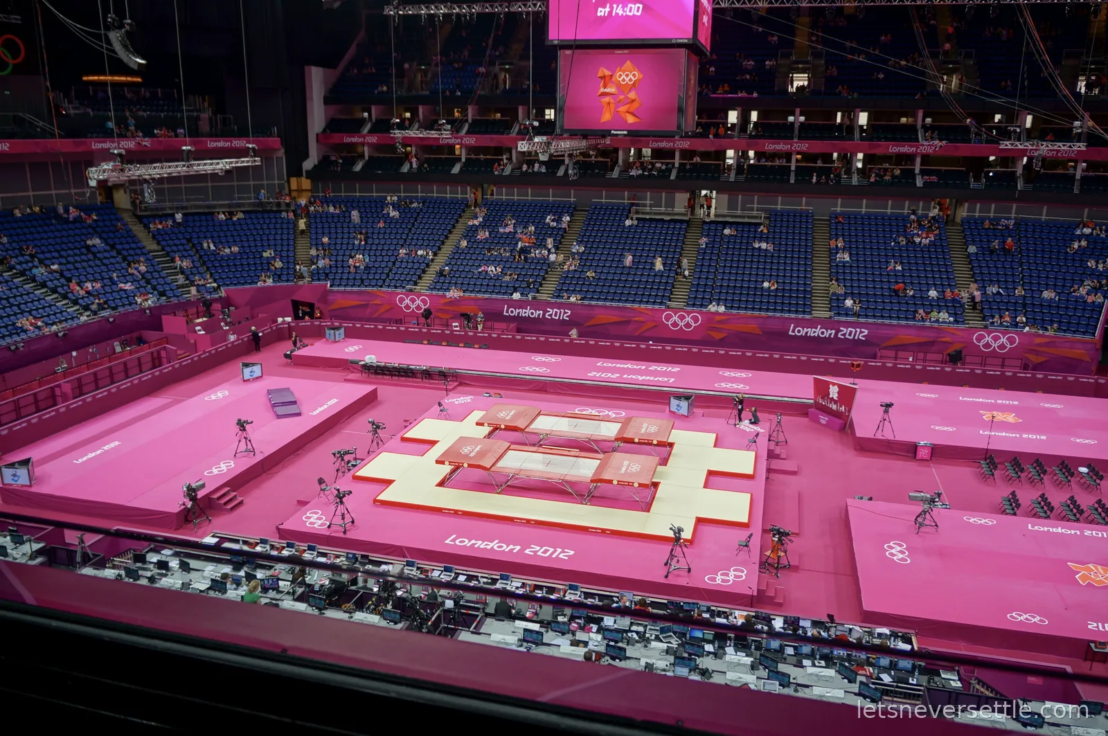 View from high in the North Greenwich Arena over the pink London 2012 field of play, two competition trampolines side by side and a long bench of press positions below
