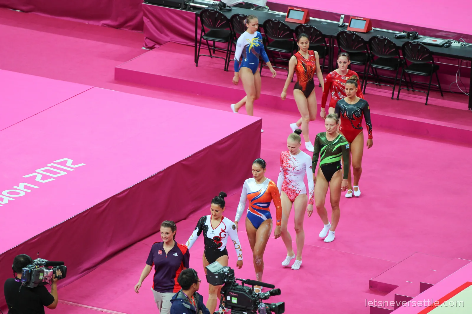 A line of women trampoline gymnasts in national leotards walking out onto the pink London 2012 floor past television cameras