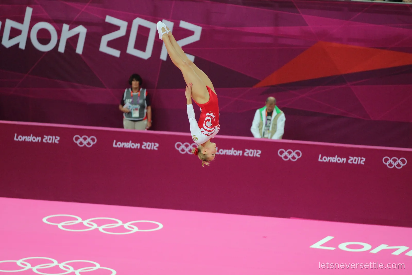 A trampoline gymnast in a red and white leotard upside down in mid air against the London 2012 branded wall of the arena