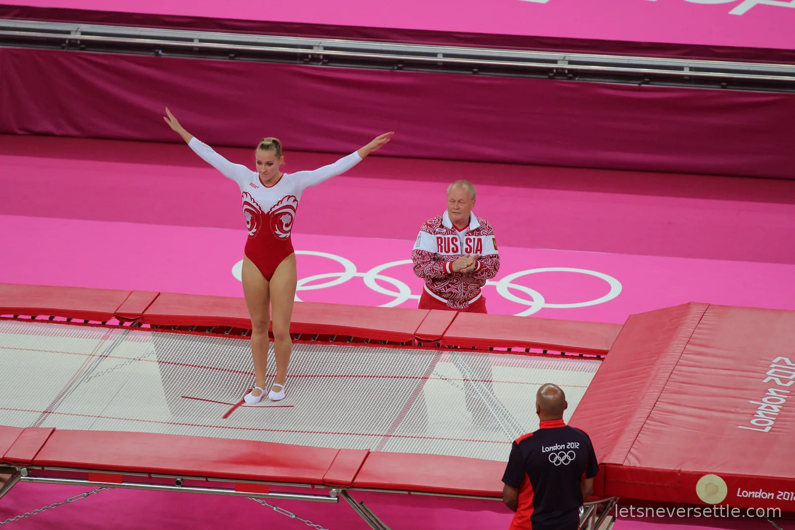 A Russian trampoline gymnast standing on the trampoline with arms raised at the end of a routine, her coach in a Russia tracksuit applauding on the pink floor behind