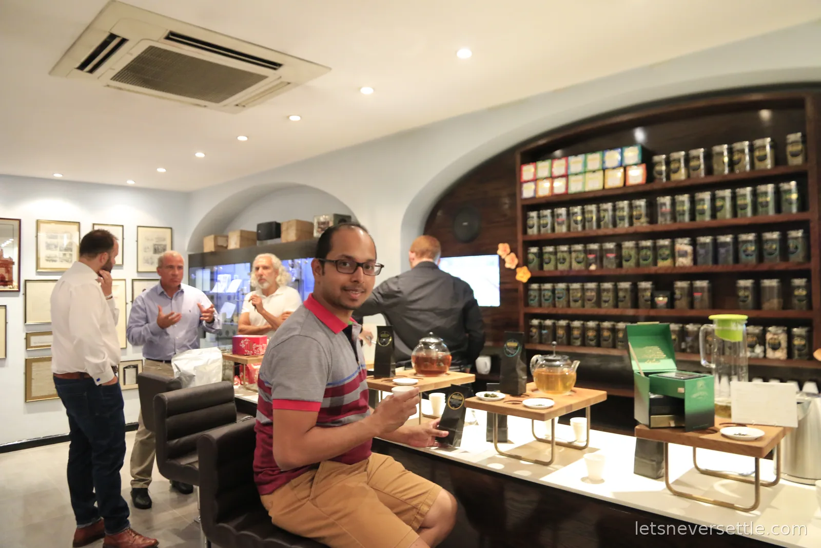 Dileep sitting at the tasting counter inside Twinings with glass teapots brewing in front of him and a wall of tea caddies behind