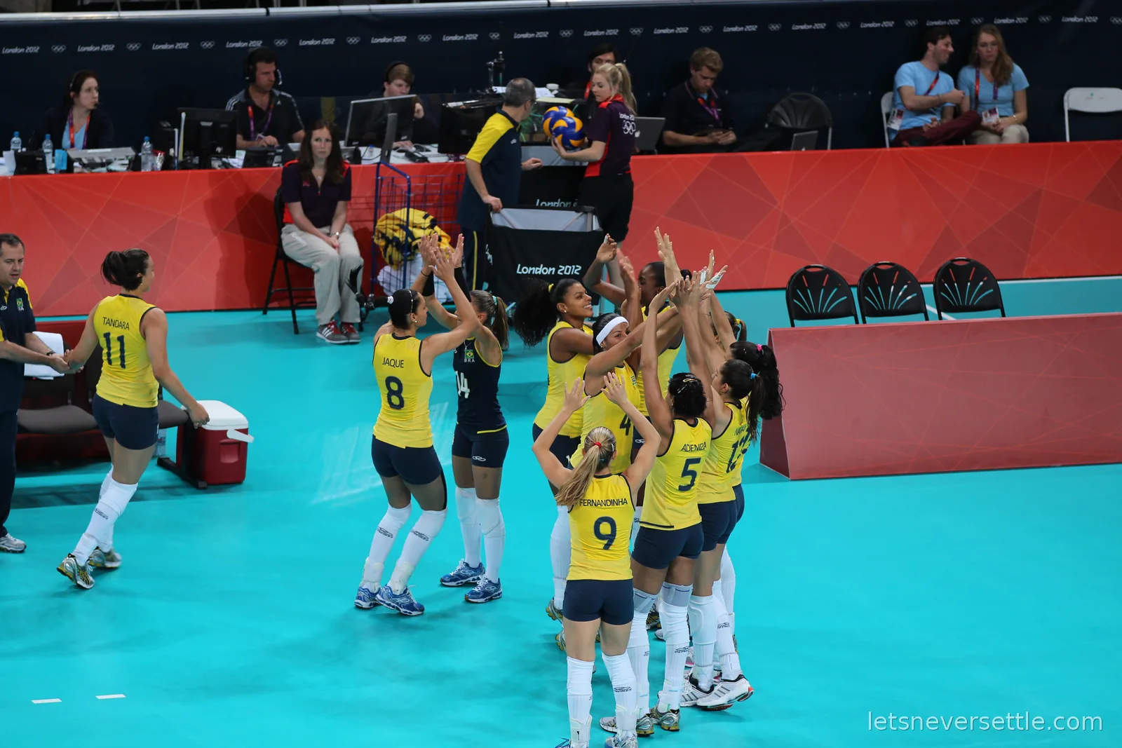 Brazilian women's volleyball players in yellow huddling and reaching for the ball courtside at London 2012