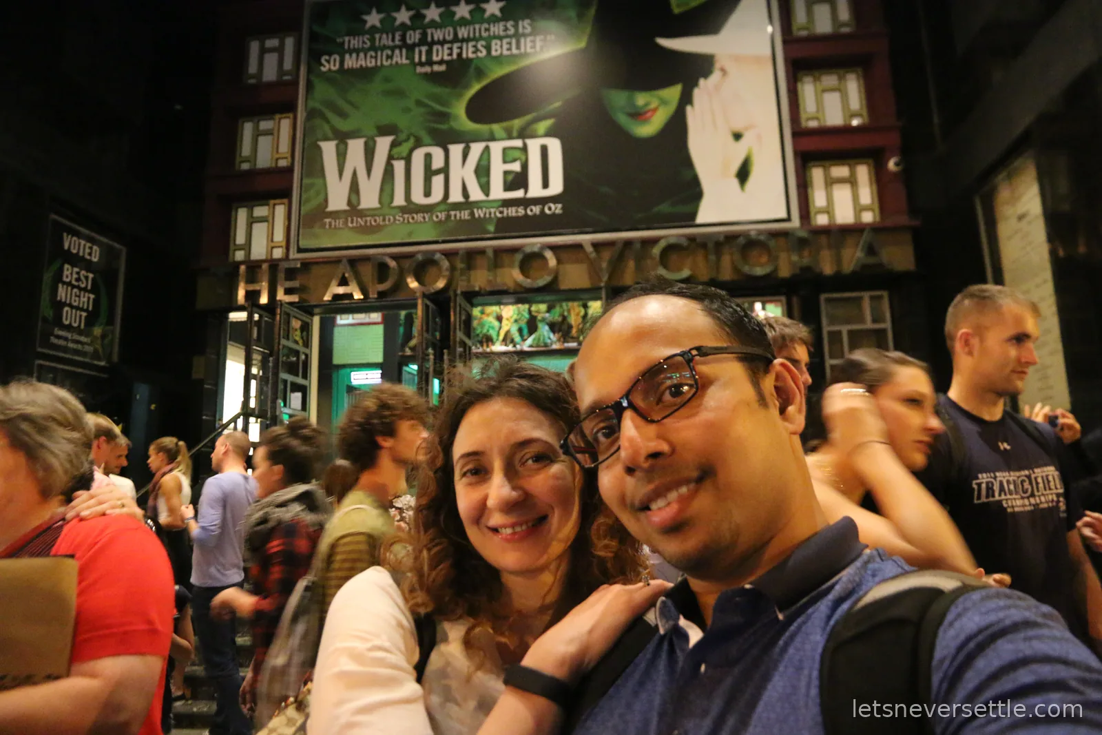 Inga and Dileep in the crowd outside the Apollo Victoria theatre at night, the illuminated Wicked sign above them