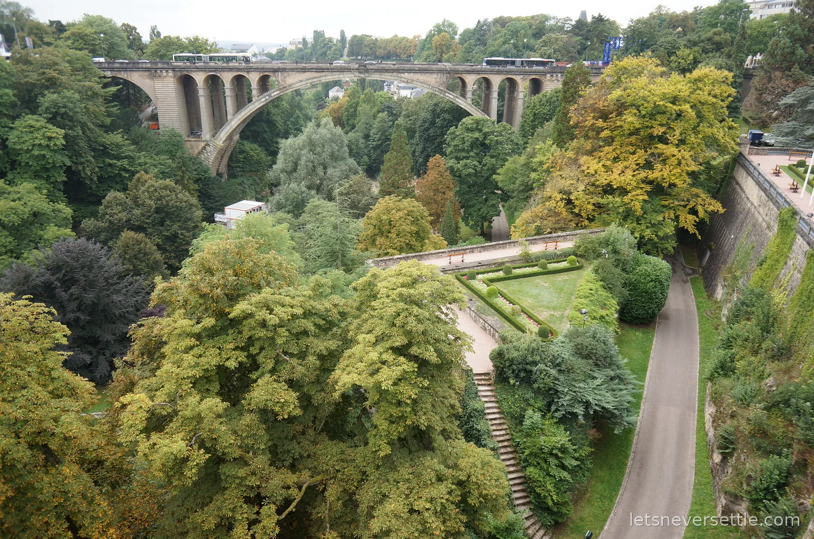The stone arch of the Adolphe Bridge spanning the leafy Petrusse valley in Luxembourg City, with buses crossing and a triangular hedged garden below