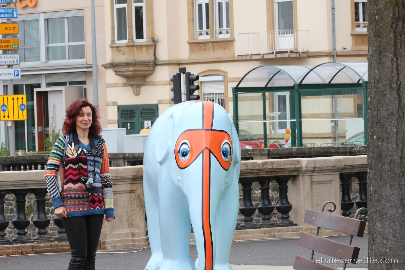 Inga standing next to a light blue painted elephant sculpture with an orange face mask design, on a stone bridge in Luxembourg City
