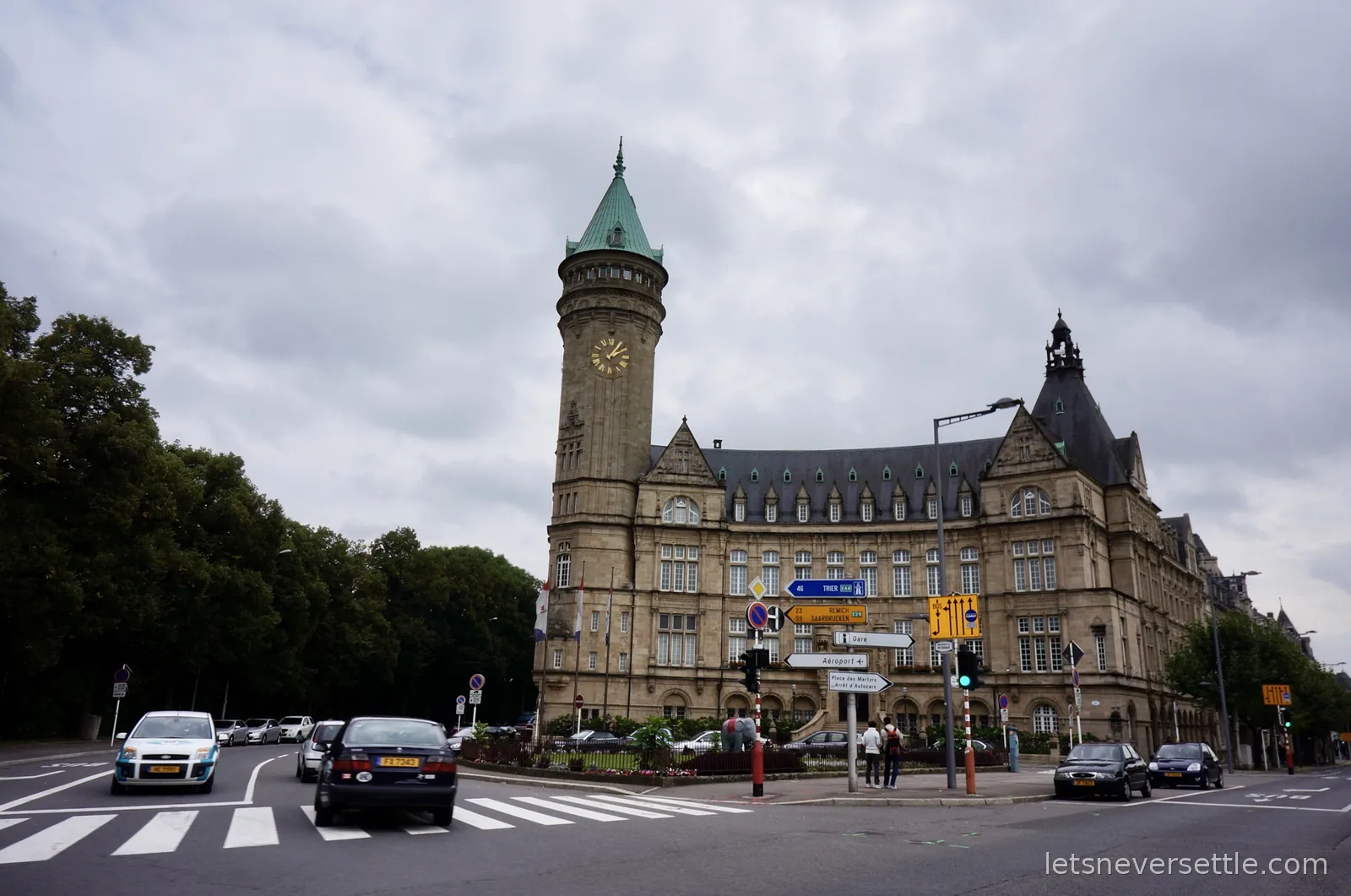 The stone State Savings Bank building in Luxembourg City with its tall clock tower and green spire, behind road signs pointing to Trier, Remich and Saarbrucken
