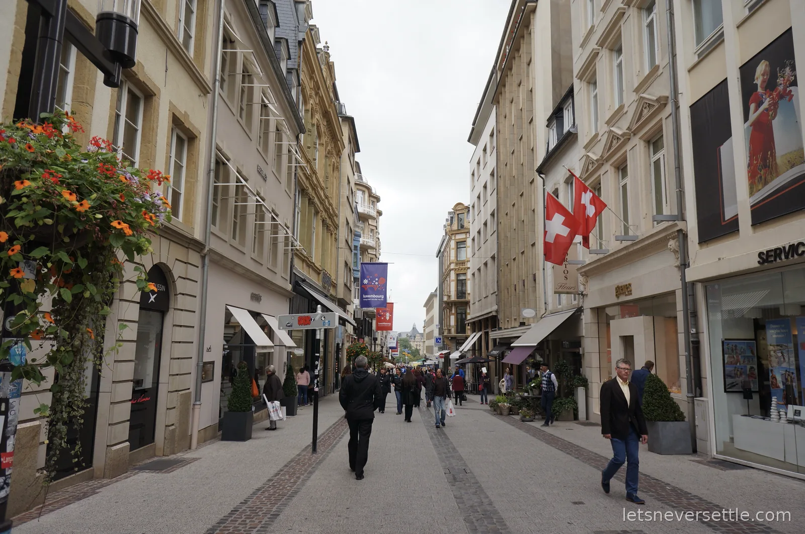 Pedestrian shopping street in Luxembourg City lined with stone facades, hanging flower baskets, shoppers and Luxembourg city banners