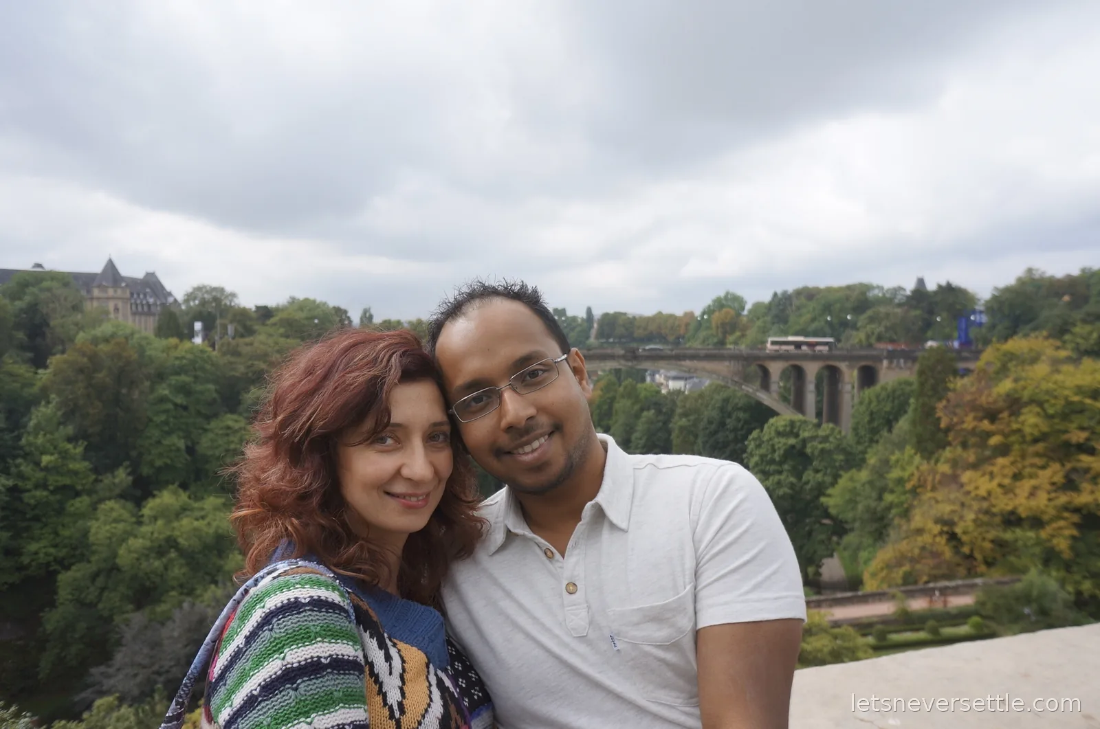 Inga and Dileep smiling together on the ramparts of Luxembourg City, with the Adolphe Bridge and wooded valley behind them