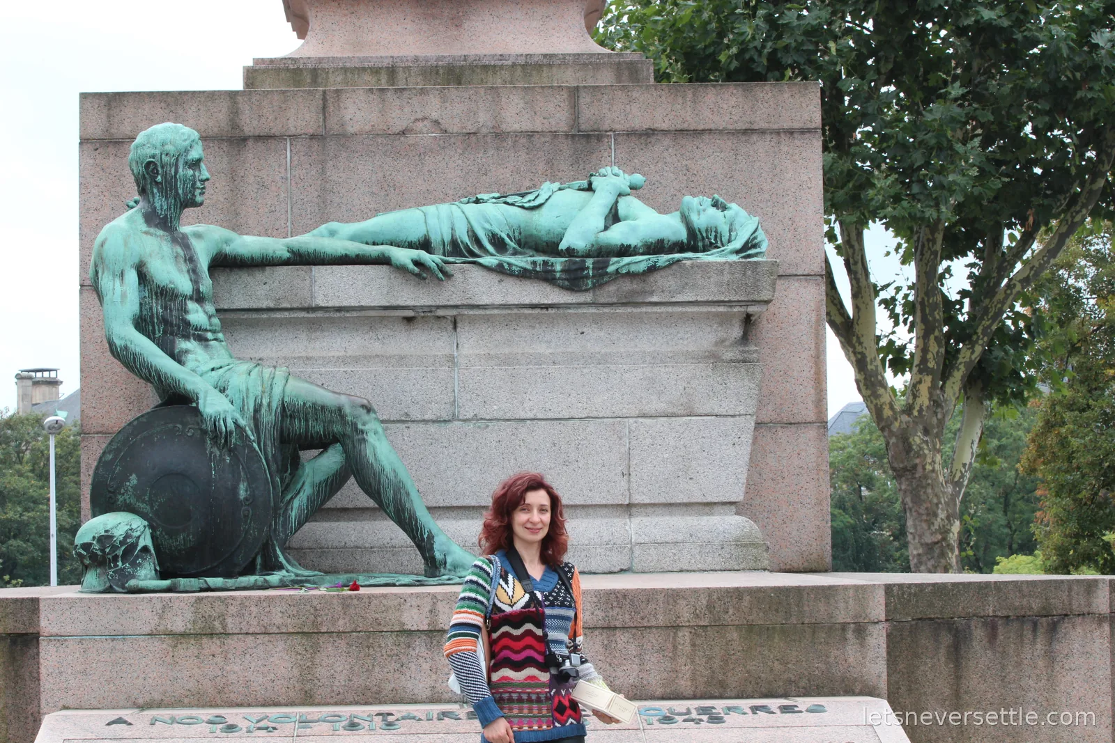 Inga standing at the base of the Monument of Remembrance in Luxembourg City, in front of green bronze figures of a seated soldier and a lying figure, above an inscription to the war volunteers of 1914 to 1918 and 1940 to 1945