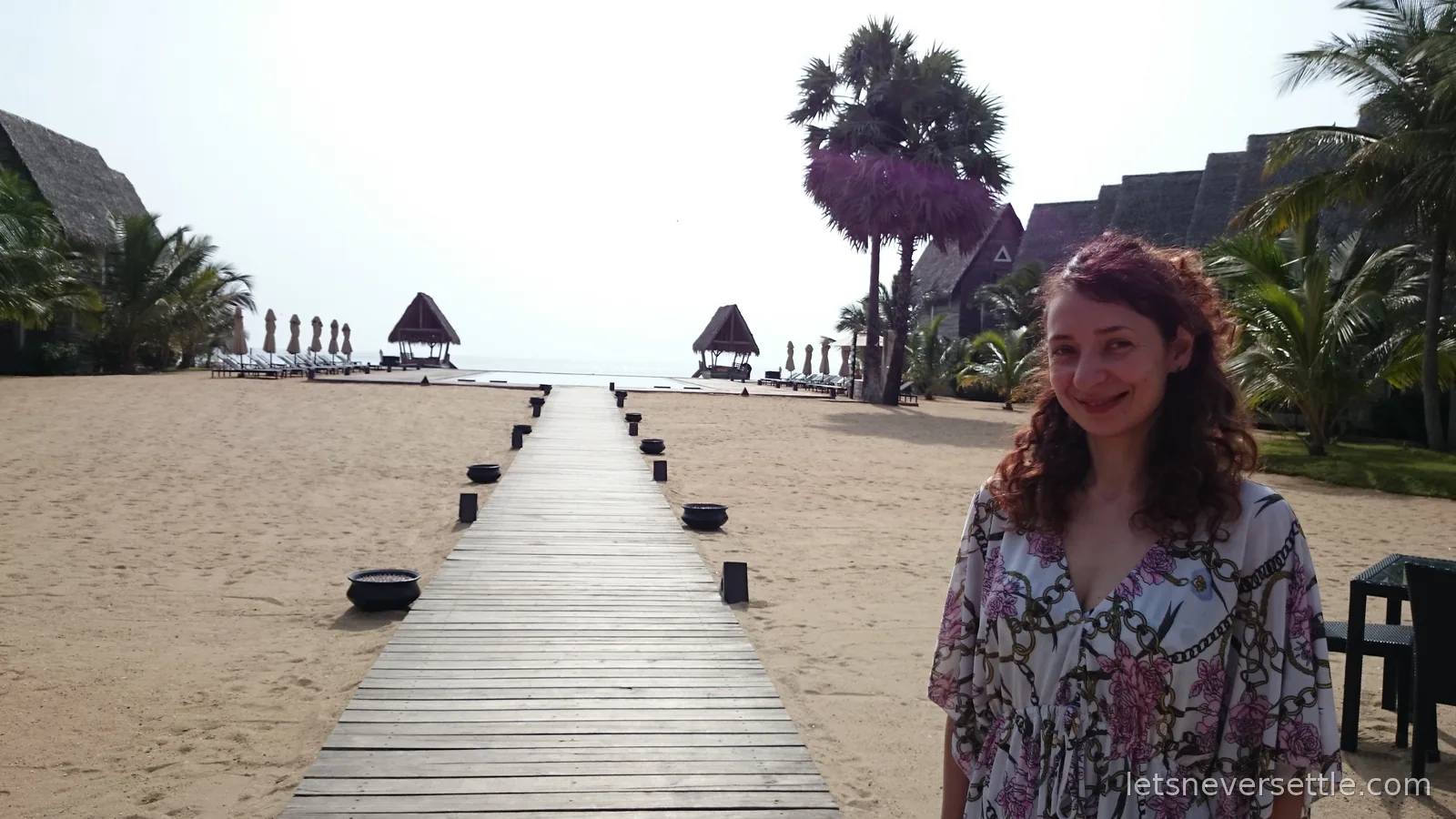 Inga smiling beside the long wooden boardwalk that runs across pale sand between palm trees to the sea