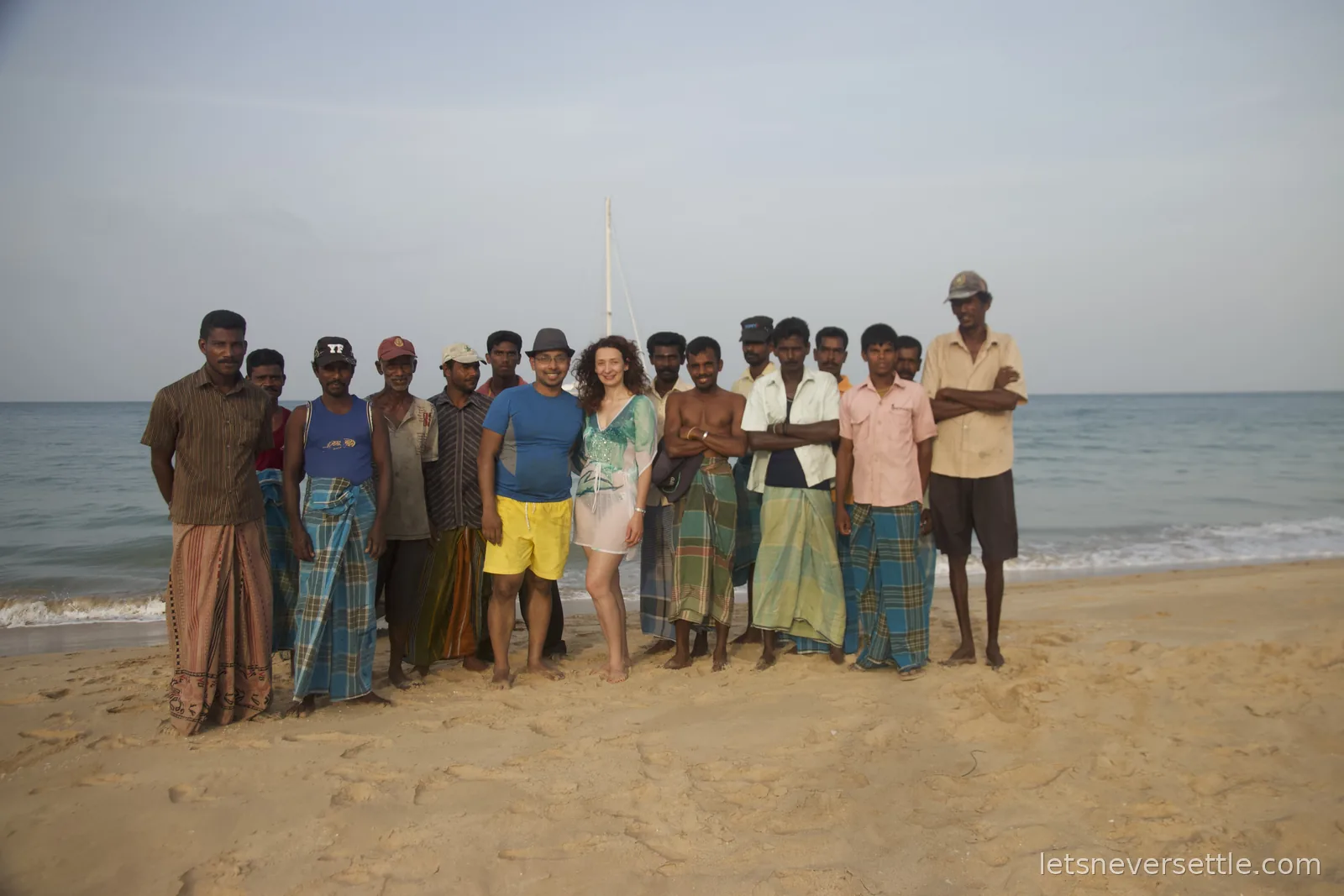 Inga and Dileep posing on the beach with a large group of local fishermen in sarongs, a catamaran mast behind