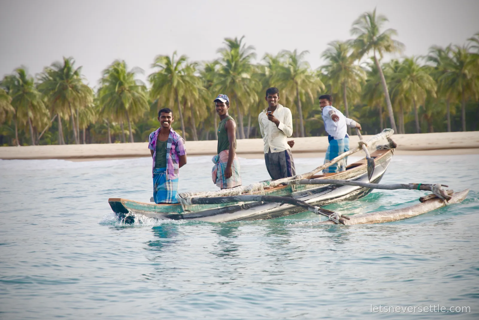 Four fishermen standing on a wooden outrigger canoe on turquoise water, coconut palms lining the beach behind