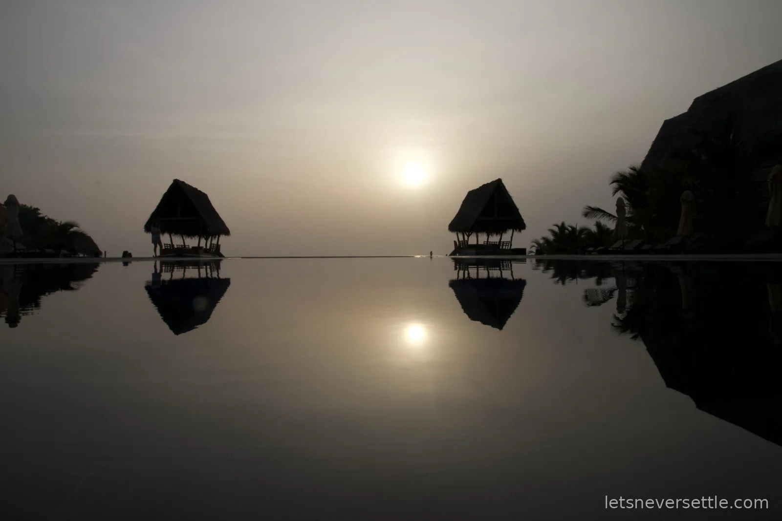 Sunrise over the sea reflected in a dark infinity pool, two thatched cabanas silhouetted on either side