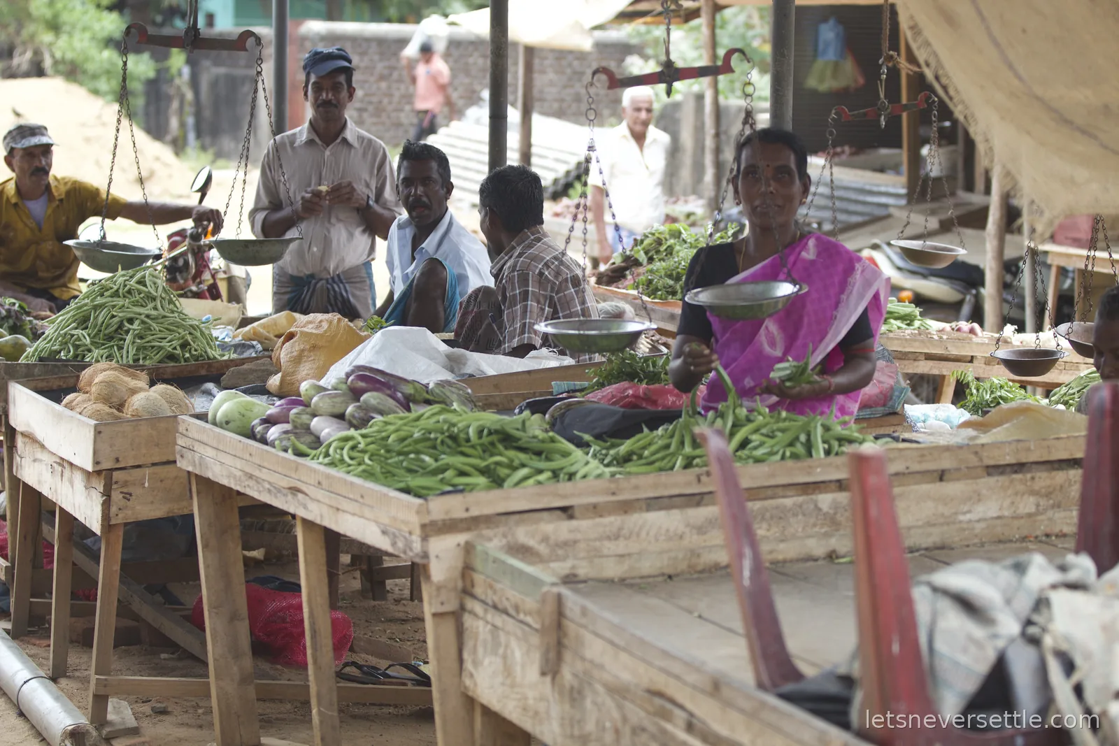 A local vegetable market with wooden tables of okra and aubergines, hanging balance scales and a stallholder in a pink sari