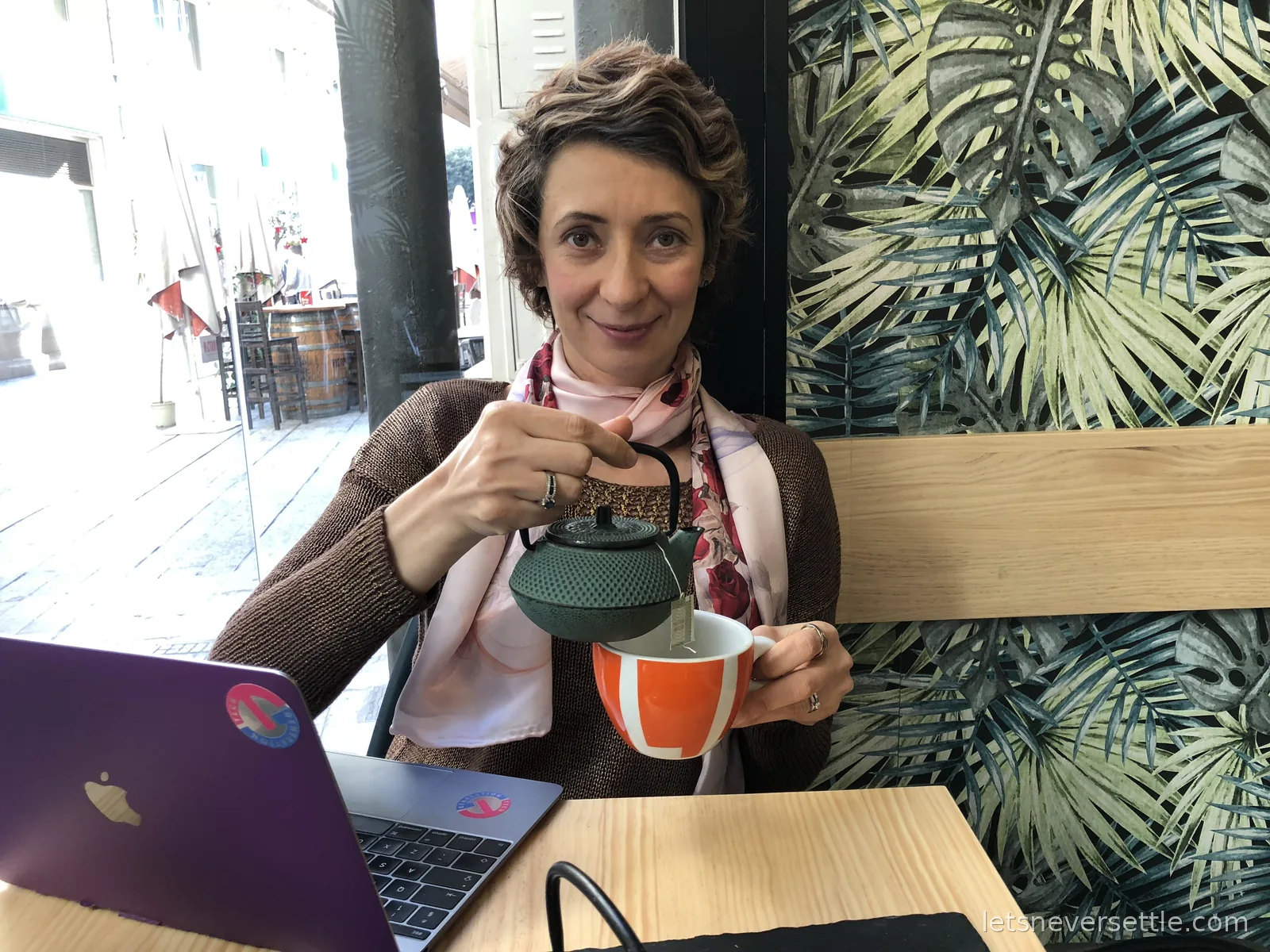 Inga pouring tea from a small green cast-iron teapot into an orange and white cup at a cafe table with a laptop, palm-print wall behind