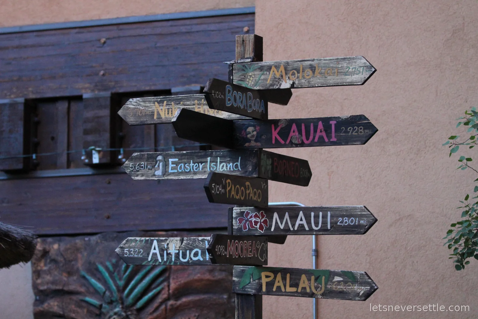 A signpost of hand painted wooden arrows pointing to Pacific islands including Kauai, Maui, Easter Island and Palau with distances in miles