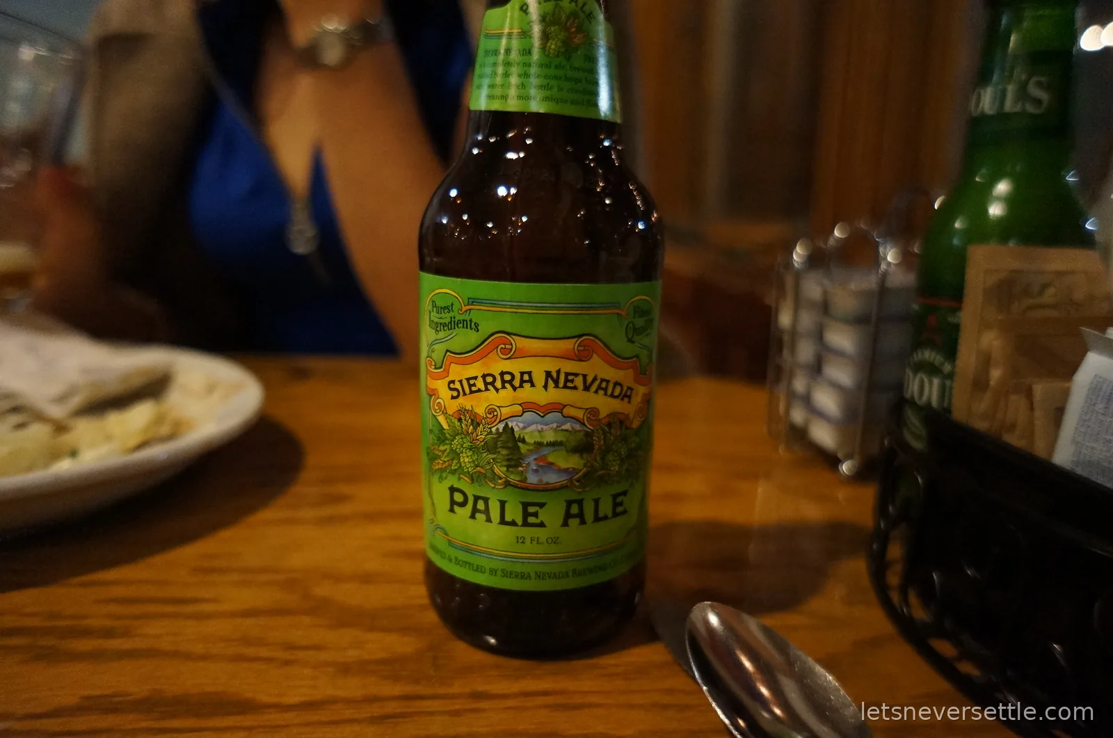 A bottle of Sierra Nevada Pale Ale on a wooden dinner table with plates and drinks behind