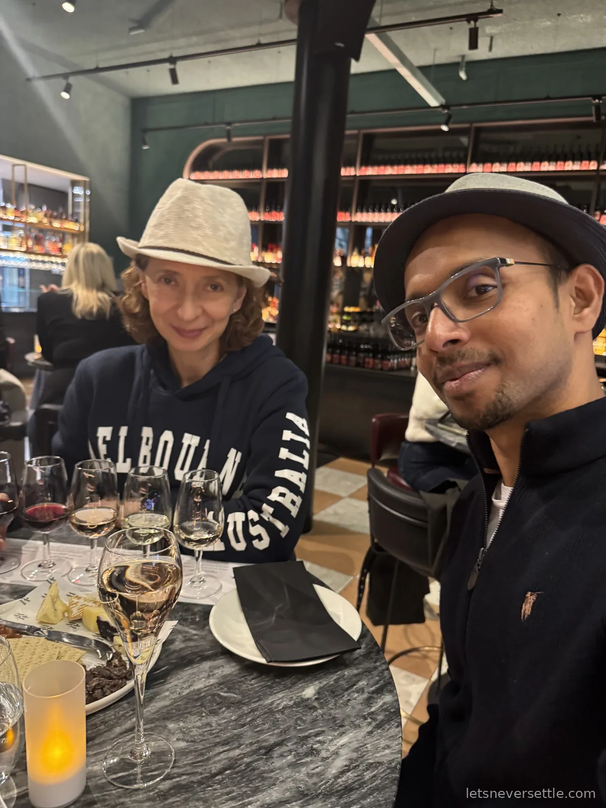 Inga and Dileep in hats at a marble bar table with a flight of red, white and sparkling wines, bottle shelves lit behind