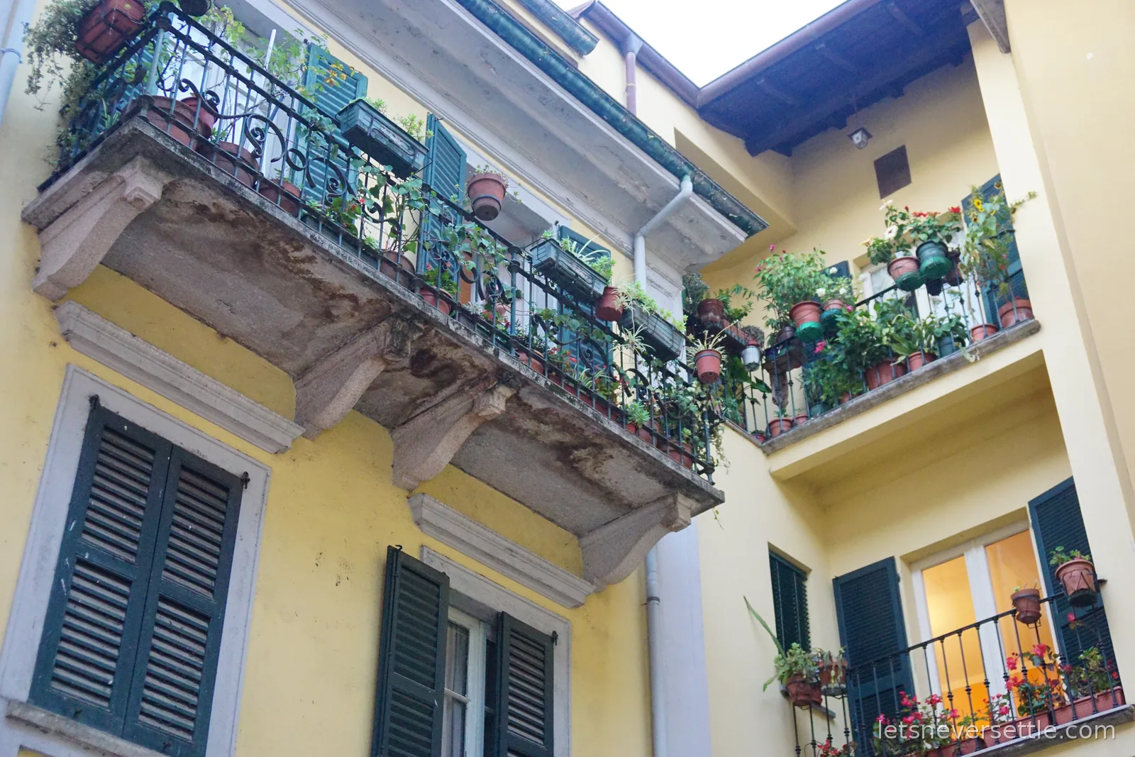 Looking up at two storeys of a yellow building with dark green shutters, its stone and wrought iron balconies crowded with pots of trailing plants and geraniums