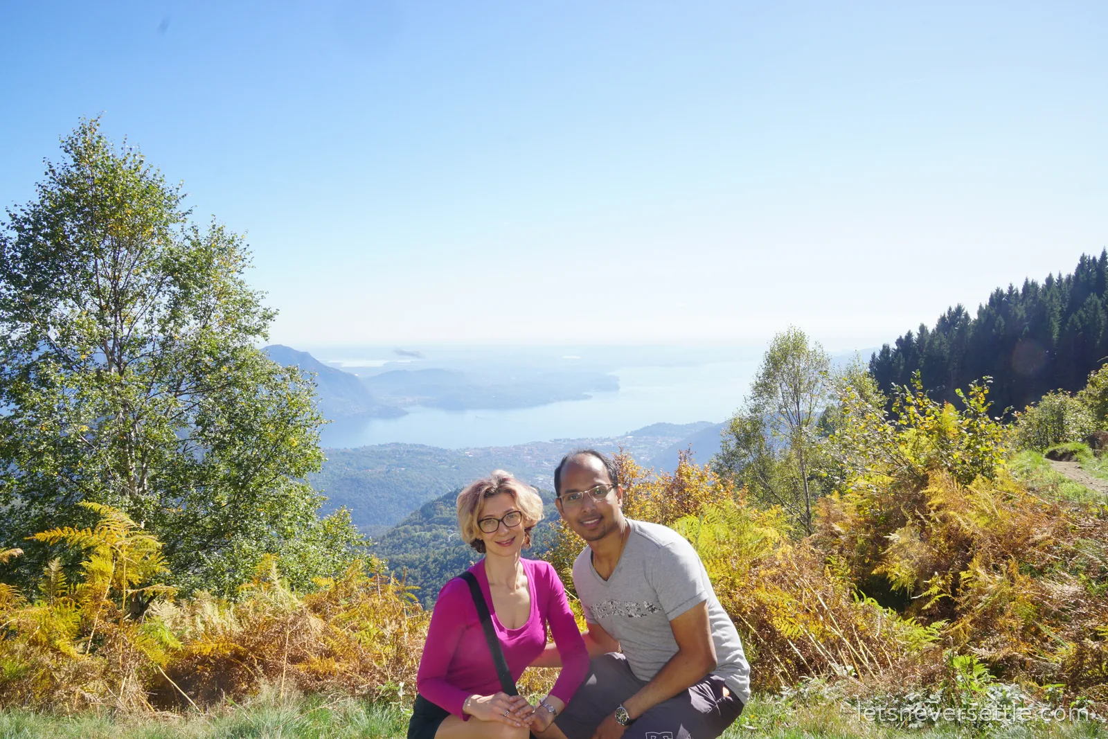 Inga and Dileep crouching together among golden bracken on a high slope, with layers of wooded ridges and Lake Maggiore in the haze behind them