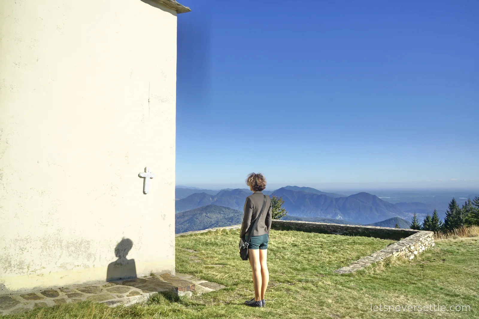 Inga standing on the grass beside the white chapel wall with her back to the camera, looking out over blue mountain ridges and a flat hazy plain beyond