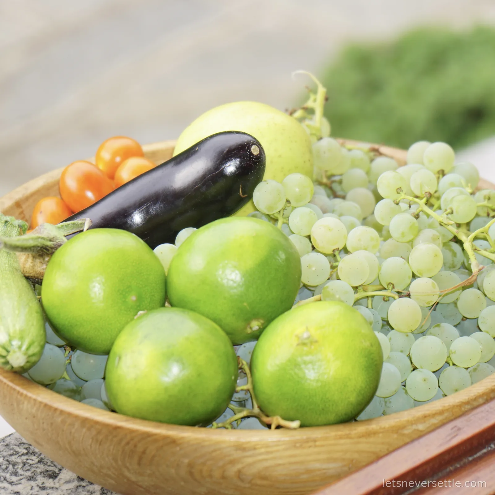 A wooden bowl on a stone table piled with white grapes on the stem, a glossy dark aubergine, a small courgette with its flower still attached, and orange and green tomatoes