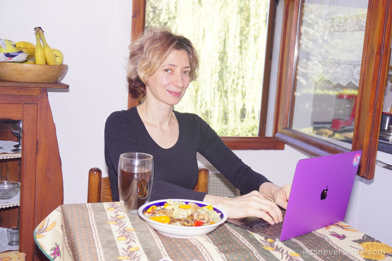 Inga sitting at a table with a floral cloth, typing on a purple laptop beside a plate of mushrooms and peppers and a tall glass, with a wooden dresser and a bowl of bananas behind her and green trees through the window