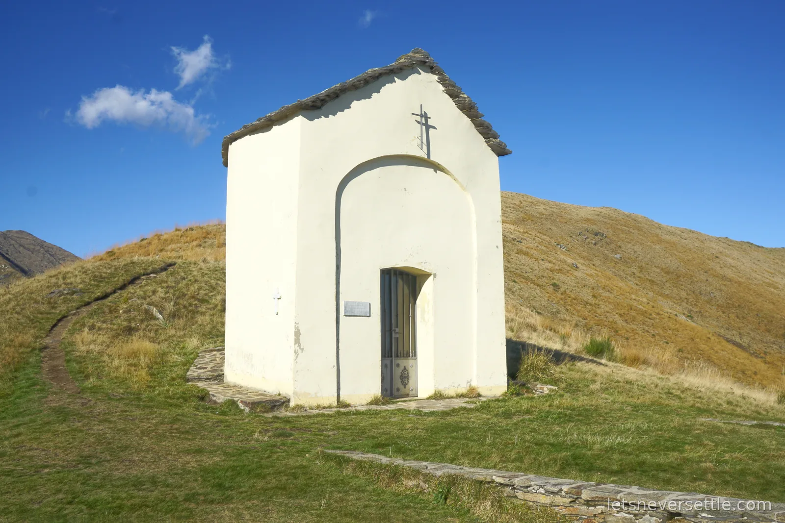 A small square white chapel with a stone slab roof and an iron cross on the gable, standing alone on a grassy ridge under a deep blue sky