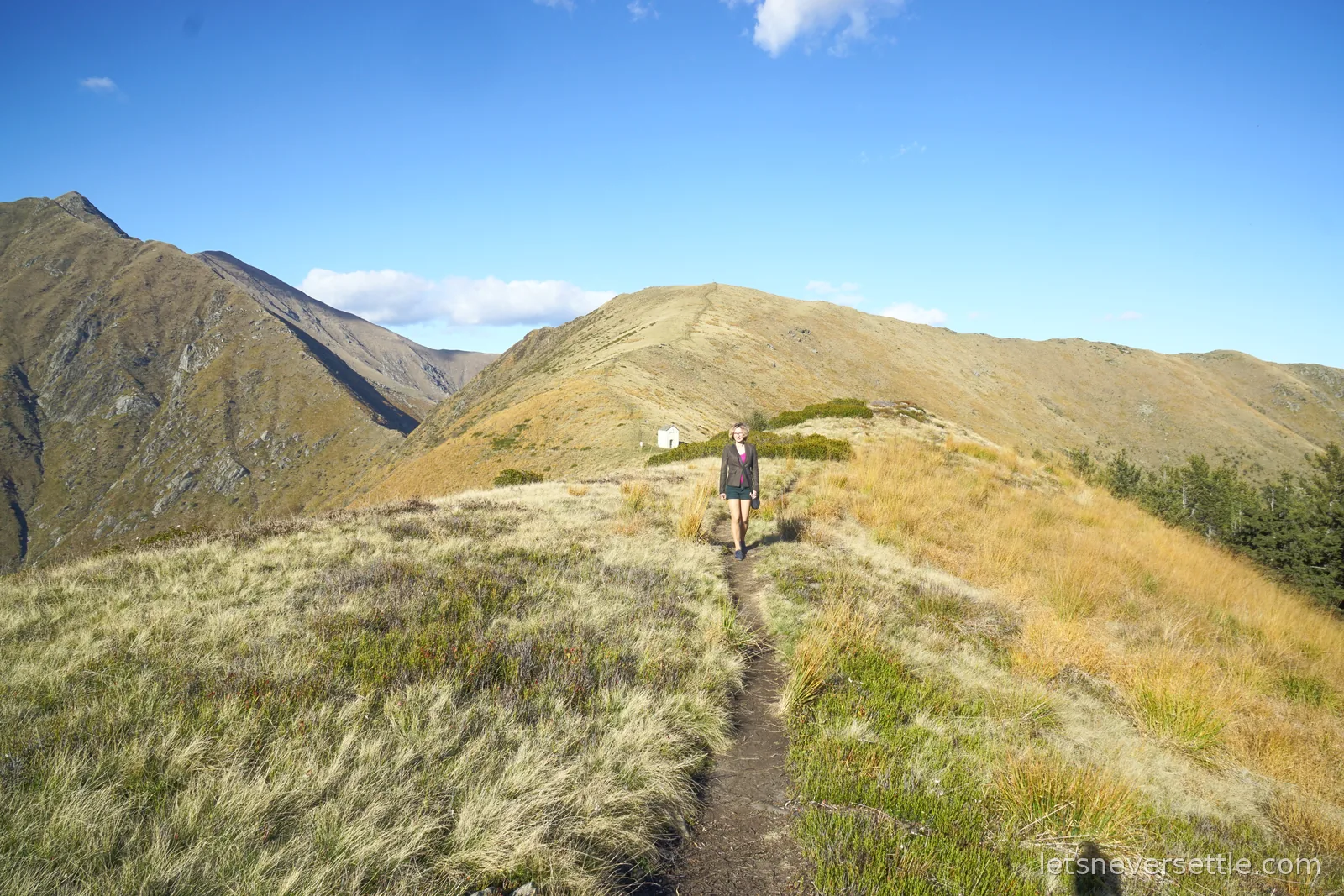 Inga walking towards the camera along a narrow earth path running the length of a grassy ridge, with the small white chapel visible far behind her and steep slopes on both sides